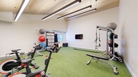 Located in Colorado Springs, CO, this dedicated fitness studio offers a thoughtfully designed workout environment ideal for a wide range of training styles. The space is defined by its striking vaulted ceiling clad in natural wood paneling, which lends warmth and architectural character while contributing to a sense of openness and height. Sleek linear pendant lights run parallel along the ceiling, providing bright, even illumination throughout the room.

The floor is covered in vibrant green turf, offering a durable, low-impact surface well-suited for both cardio and functional training activities. The crisp white walls keep the environment feeling clean and energizing, complemented by a wall-mounted flat-screen television for entertainment or guided workouts.

The studio is outfitted with an impressive array of equipment, including multiple stationary spin bikes, a rowing machine, a multi-functional training rack with TRX suspension straps, resistance bands, and battle ropes. A dedicated storage rack houses an assortment of kettlebells, medicine balls, and stability balls of varying sizes, keeping the space organized and efficient. A weight bench adds further versatility for strength training exercises.

Natural light filters in through a window, enhancing the overall ambiance. Together, these features create a fully functional, private fitness studio designed to support diverse workout routines.