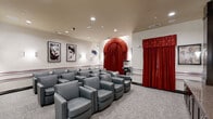 Located at 900 Grand Central Dr in Hamilton, NJ, this dedicated screening room is designed with a classic Hollywood cinema theme that delivers a immersive, boutique movie-watching experience. The single-floor space is thoughtfully arranged with rows of plush, grey leather club chairs tiered for optimal sightlines toward a large flat-screen display mounted within a sleek, dark-wood built-in entertainment console. Recessed ceiling lighting and elegant wall sconces provide warm, ambient illumination throughout, complementing the room's refined atmosphere. A dramatic arched alcove framed by rich red curtains serves as a striking focal point, while matching red drapery panels along the adjacent wall reinforce the theatrical ambiance. The walls are adorned with framed vintage Hollywood portraits and iconic movie posters — including likenesses of Marilyn Monroe, Audrey Hepburn, The Godfather, and King Kong — adding a curated, cinematic character to the space. A decorative film reel wall sculpture adds an additional nod to classic cinema culture. A popcorn cart stationed near the seating area completes the authentic movie-going experience. Neutral carpet flooring, a red accent stripe along the wainscoting, and recessed ceiling speakers round out this well-appointed screening room.