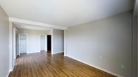 This single-floor space offers a clean, modern aesthetic defined by warm hardwood-style flooring and a neutral, light grey colour palette throughout. The main living area is generously proportioned, providing an open and airy feel that lends itself well to a variety of furniture arrangements. The textured white ceilings add a classic finishing touch, while crisp white baseboards provide a sharp contrast to the rich tones of the flooring below.

A structural beam detail subtly separates the main living area from a secondary space beyond, which appears to lead into additional rooms. Visible through the open passageway is a room with a built-in closet featuring bi-fold doors, suggesting a bedroom with practical storage. A dark-toned door further along the hallway adds depth and visual interest to the layout.

The overall ambiance is bright and neutral, making the space highly adaptable for personalization. The combination of durable flooring, clean wall finishes, and a thoughtful layout creates a functional and welcoming environment. The absence of clutter and the simplicity of the finishes make this a versatile space suitable for a range of uses.
