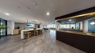 Located at 375 Camino De La Reina in San Diego, California, this single-floor communal space offers a thoughtfully designed environment that blends comfort, functionality, and contemporary style. The open layout is anchored by large-format tile flooring throughout, lending a clean and cohesive aesthetic across all zones.

At the heart of the space is a well-appointed kitchen and bar area featuring a white quartz island with bar seating, light wood cabinetry, a textured terrazzo backsplash, and a wall-mounted display screen — ideal for casual gatherings or informal meetings. Adjacent to this is a communal dining area with a long natural wood table, upholstered seating, and curated wall art that adds a vibrant, creative touch.

The lounge area is generously proportioned, furnished with deep navy sofas, layered rugs, and an assortment of coffee tables in marble and warm-toned wood. Floor-to-ceiling glass walls flood the space with natural light and frame views of the surrounding outdoor greenery, creating a seamless indoor-outdoor connection.

A more intimate zone features a private booth enclosure with teal upholstered banquette seating, warm brass accents, and wood paneling — perfect for quiet conversation or focused work. Dedicated desk space with task seating is also available along the perimeter. Together, these areas create a versatile, welcoming environment suited to both social and professional use.