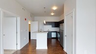 This single-floor space showcases a thoughtfully designed open-concept layout centered around a contemporary kitchen. The kitchen is outfitted with dark espresso-toned cabinetry that contrasts sharply against a classic white subway tile backsplash, creating a bold and stylish aesthetic. Stainless steel appliances, including a refrigerator, range, and dishwasher, contribute a polished, modern finish throughout the space.

A central kitchen island with a granite countertop and crisp white base serves as a functional focal point, ideal for meal preparation or casual dining. The rich, dark hardwood-style flooring extends throughout the open area, adding warmth and continuity to the space.

Pendant lighting fixtures hang above the kitchen area, providing targeted illumination that complements the recessed ceiling lights. The neutral white walls and standard ceiling height create a clean, airy atmosphere. A hallway visible beyond the kitchen suggests additional rooms, while a closed door near the kitchen hints at extra storage or utility space. Overall, the unit presents a well-appointed, functional living environment with modern finishes.