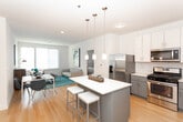 Located at 22 E 19th St in Bayonne, NJ, this single-floor residence offers a thoughtfully designed open-concept layout with modern finishes throughout. The kitchen is a standout feature, equipped with stainless steel appliances including a gas range, over-the-range microwave, dishwasher, and refrigerator. Two-tone cabinetry pairs gray lower cabinets with white upper cabinets, complemented by a classic subway tile backsplash and a central island with pendant lighting overhead. Light hardwood flooring extends through the kitchen and into the adjacent dining and living areas, creating a cohesive, airy feel. The living room benefits from large windows that bring in generous natural light and offer views of the surrounding Bayonne neighborhood. The bedrooms feature soft carpeting, neutral walls, and well-sized windows that maintain a calm, restful atmosphere. The bathroom is finished with large-format tile flooring, a gray shaker-style vanity with a white countertop, a wide mirror with overhead lighting, and a full tub and shower combination. Recessed lighting and clean architectural lines contribute to the unit's overall contemporary and functional character.