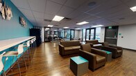 This single-floor commercial space is thoughtfully designed to balance collaborative lounging with focused work, offering a welcoming and functional environment. Rich hardwood-style flooring runs throughout, complementing a neutral drop-tile ceiling with recessed panel lighting that keeps the space bright and well-illuminated.

The main lounge area is a standout feature, furnished with plush brown leather sofas and armchairs arranged around teal cube accent tables — a design choice that adds a pop of color echoed in the bold teal accent walls. Decorative wall art and built-in counter seating along the teal-painted wall add character and utility, while vending machines nearby provide convenient amenities.

A dedicated dining zone features round tables with sleek metal-frame chairs, creating an informal gathering spot ideal for breaks or casual meetings. Glass-partitioned walls border a separate workspace area equipped with individual workstations, offering visual connectivity while maintaining a quieter environment for focused tasks.

Large floor-to-ceiling windows and glazed entry doors flood the space with natural light and provide views to the exterior. A reception or service counter with an overhead flat-screen display anchors one end of the space, adding a professional touch. The overall ambiance is modern, clean, and purposefully designed for comfort and productivity.