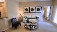 This single-floor space presents a thoughtfully designed living room that combines comfort with contemporary style. The living area is anchored by a crisp white sofa adorned with green and black accent pillows, complemented by a sleek black armchair that adds contrast to the neutral color palette. A round glass coffee table with a metal frame serves as a functional centerpiece, displaying decorative accents that enhance the room's aesthetic. The walls are finished in warm beige tones, creating an inviting atmosphere, while plush wall-to-wall carpeting adds warmth underfoot. A trio of framed botanical prints mounted above the sofa introduces a natural element to the décor, tying together the green accents throughout the space. Soft lighting is provided by a classic table lamp positioned on a mirrored side table, casting a warm glow across the room. A hallway visible in the background suggests additional rooms beyond the living area. The overall ambiance is calm, curated, and welcoming, making it a comfortable space for both relaxation and everyday living.