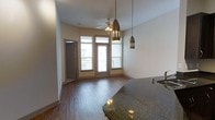 This single-floor residence showcases an open-concept living and kitchen area designed for both functionality and style. The kitchen features dark granite countertops that provide a striking visual anchor, complemented by sleek metallic pendant lights that hang at varying heights, adding a modern, decorative touch to the space.

The living area extends naturally from the kitchen, unified by warm hardwood-style flooring that runs throughout. Tall ceilings enhance the sense of openness and volume, while a ceiling fan with an integrated light fixture provides both practical airflow and ambient illumination.

Natural light is a defining characteristic of this space, with large glass-paned doors and flanking windows along the far wall drawing in generous amounts of daylight. These doors open directly onto a private balcony, offering outdoor access and views of the surrounding area.

A secondary door on the adjacent wall provides additional access or leads to a utility space. The neutral wall tones throughout create a clean, versatile backdrop that complements the warm flooring and rich countertop finishes. Overall, the space presents a well-balanced combination of modern fixtures and inviting design elements.