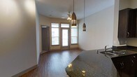 This single-floor residence showcases an open-concept living and kitchen area designed for both functionality and style. The kitchen features dark granite countertops that provide a striking visual anchor, complemented by sleek metallic pendant lights that hang at varying heights, adding a modern, decorative touch to the space.

The living area extends naturally from the kitchen, unified by warm hardwood-style flooring that runs throughout. Tall ceilings enhance the sense of openness and volume, while a ceiling fan with an integrated light fixture provides both practical airflow and ambient illumination.

Natural light is a defining characteristic of this space, with large glass-paned doors and flanking windows along the far wall drawing in generous amounts of daylight. These doors open directly onto a private balcony, offering outdoor access and views of the surrounding area.

A secondary door on the adjacent wall provides additional access or leads to a utility space. The neutral wall tones throughout create a clean, versatile backdrop that complements the warm flooring and rich countertop finishes. Overall, the space presents a well-balanced combination of modern fixtures and inviting design elements.