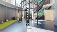 Located at 575 Princess St in Kingston, Ontario, this fitness facility offers a comprehensive and thoughtfully designed workout environment across a single expansive floor. The space features an industrial-style aesthetic, with exposed ductwork and structural elements painted in dark tones overhead, complemented by bright linear fluorescent lighting that ensures the entire floor is well-lit throughout.

The facility is organized into distinct training zones. A dedicated cardio area houses a row of stationary spin bikes and treadmills, positioned alongside interior windows and a wall-mounted display screen. Adjacent to this is an open functional training zone featuring large mirrors along the wall, a green turf section, and a functional rig equipped with cable systems and weight stacks — ideal for strength and conditioning work. A vibrant graffiti-style mural adds a bold visual accent near the rig area, alongside a staircase with an industrial metal railing.

The flooring transitions between polished concrete and rubber matting across the various zones, providing both durability and appropriate surface support. Sage green and warm terracotta accent walls add color and visual variety throughout the space. The overall ambiance is energetic and modern, combining urban industrial design with a fully functional, multi-purpose fitness layout suited for a wide range of training styles.