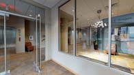 This well-appointed single-floor commercial space blends modern design with functional amenities, creating a professional yet welcoming environment. The entry corridor sets an elegant tone with floor-to-ceiling glass partitions, chrome hardware, and a decorative pendant chandelier visible through the glazing, leading guests into a bright, open interior.

The central gathering area is a standout feature, anchored by a sleek linear gas fireplace set into a refined tile surround with a large-format display mounted above. Comfortable leather seating and a coffee table arrangement create an inviting lounge zone, accented by warm hardwood flooring that extends throughout the main areas. Curated art pieces and decorative accents add subtle personality to the space.

Adjacent to the lounge is a generously sized open dining and breakroom area, outfitted with multiple tables and mixed seating options. A fully equipped kitchen runs along one side, featuring stainless steel appliances, a built-in microwave, bar-height seating, and ample cabinetry with a dark tile backsplash. A separate windowed dining alcove offers a bright, sunlit area for more intimate seating, with natural light filtering through horizontal blinds that overlook the surrounding exterior.

Throughout, recessed ceiling lighting provides consistent illumination, complementing the clean, neutral palette of warm grays, taupes, and natural wood tones that define the overall aesthetic.