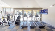 This fitness center is designed to accommodate a comprehensive range of workout routines within a single, open floor plan. The space is divided into distinct functional zones, each outfitted with purpose-specific equipment. The cardio area features a row of treadmills and a stair climber machine positioned along a wall of large windows, allowing natural light to fill the space while offering outdoor views. A wall-mounted flat-screen television provides entertainment during cardio sessions.

The strength training section is well-appointed with a full dumbbell rack ranging across multiple weight increments, a cable machine, and various resistance equipment. Floor-to-ceiling mirrors line the walls in this area, enhancing the sense of space and allowing users to monitor their form. Overhead, a combination of recessed and pendant lighting ensures consistent illumination throughout.

A functional training zone features adjustable benches, medicine balls, stability balls, and accessory racks, supporting flexibility and cross-training workouts. Structural columns articulate the layout, and glass-paneled interior partitions provide visual connectivity between areas. The durable checkerboard rubber flooring throughout offers both safety and a clean, professional aesthetic.