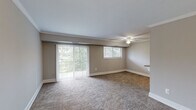 This single-floor, 1-bedroom, 1-bathroom unit offers a thoughtfully organized layout across a compact yet efficiently utilized footprint. The living room serves as the central gathering space, featuring warm neutral-toned carpet, crown molding, a ceiling fan with light fixture, and a large sliding glass door flanked by vertical blinds that opens to an outdoor balcony. A secondary window allows additional natural light and greenery views, creating a bright and welcoming ambiance.

The galley-style kitchen is positioned off the living area and is well-equipped with warm wood cabinetry, granite-style countertops with a breakfast bar overhang, track lighting, a gas range, and a black refrigerator. Light wood-look flooring lines the kitchen floor, providing a clean contrast to the surrounding carpeted rooms.

The bedroom is a generously sized private retreat with its own ceiling fan, a wide window with blinds offering natural light and outdoor views, and plush carpeting throughout. A dedicated closet provides practical storage, while a hallway connects the main areas of the unit. The bathroom features a full tub-shower combination with white subway tile surround, a vanity with sink, and a mirrored medicine cabinet, maintaining a clean and classic aesthetic throughout.