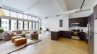 This single-floor urban loft combines industrial character with modern finishes throughout an open, airy layout. Soaring ceilings with exposed ductwork lend an authentic loft aesthetic, while polished concrete floors unify the main living areas. A wall of large, steel-framed industrial windows floods the space with natural light and frames compelling views of the surrounding cityscape.

The open-plan living area flows seamlessly from a generous lounge space to a dedicated work nook, making it highly functional. The kitchen is well-appointed with dark espresso cabinetry, stainless steel appliances including a refrigerator, range, microwave, and dishwasher, complemented by a classic white subway tile backsplash and light-toned countertops. A peninsula with bar seating serves as a natural divider between the kitchen and living areas.

The bedroom offers a quieter retreat, featuring warm hardwood flooring, bright white walls, tall ceilings, and ample closet storage. The bathroom is finished with subway tile in the tub surround, a dark-cabinetry vanity, and the same cork-toned flooring seen throughout. The overall ambiance is contemporary and cosmopolitan, with an industrial edge that is both stylish and livable.