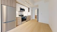 This efficiently designed one-bedroom, one-bathroom unit showcases a clean, modern aesthetic throughout its thoughtfully laid-out floor plan. Light hardwood flooring flows consistently across the main living areas, complementing the bright white walls and creating a cohesive, airy feel.

The open kitchen is well-appointed with a full suite of stainless steel appliances — including a refrigerator, electric range, over-the-range microwave, and dishwasher — paired with two-tone cabinetry featuring white upper cabinets and warm wood-grain lower cabinets. A white tile backsplash and track lighting overhead complete the modern kitchen aesthetic.

The living area is open and bright, benefiting from an unobstructed layout. The bedroom is a cozy yet functional space bathed in natural light from large floor-to-ceiling windows that frame urban views of the surrounding cityscape, adding a dynamic backdrop to the room.

The bathroom features a soaking tub with full-height white tile surround, a floating wood-grain vanity with a white countertop, and neutral tile flooring — all contributing to a clean, spa-like atmosphere.

A staircase with warm wooden treads and a natural wood handrail, positioned alongside large windows, adds architectural character and connects the unit to its entry level. Overall, the space offers a streamlined, contemporary living experience with quality finishes throughout.
