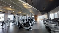 This fitness center presents a thoughtfully designed workout environment that balances functionality with modern aesthetics. The space is defined by its striking curved ceiling panels finished in a reflective material, complemented by recessed lighting that ensures consistent, even illumination throughout. The dark rubberized flooring provides both safety and a polished look, contrasting neatly with the lighter architectural elements above.

The cardio zone is prominently featured, housing a row of elliptical trainers and additional aerobic equipment arranged in an organized, accessible layout. Toward the rear, a dedicated strength and resistance training area is visible, equipped with cable machines and multi-use stations. Floor-to-ceiling windows along one wall flood the space with natural light while offering expansive city views, enhancing the overall workout experience.

Structural columns are integrated seamlessly into the design, and a curved accent wall featuring the facility's branding adds a distinct visual focal point. The overall ambiance is clean, professional, and contemporary, reflecting a high-standard fitness facility suited for a range of exercise routines.