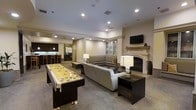 This single-floor amenity lounge is designed for comfort and entertainment, offering a well-appointed space that balances recreation and relaxation. The main lounge area is anchored by a large L-shaped sectional sofa in neutral gray, complemented by contemporary side tables and elegant table lamps that create a warm, inviting ambiance. A classic white-mantled fireplace serves as a focal point on one wall, topped by a wall-mounted flat-screen television — ideal for gatherings. Recessed lighting throughout the space provides consistent, well-distributed illumination across the carpeted floors and smooth, light-toned walls. A full-size shuffleboard table occupies a prominent position in the center of the room, making it a standout recreational feature for guests and residents alike. Toward the rear of the space, a sleek wet bar area with dark cabinetry and pendant lighting offers counter seating on bar stools, suggesting a well-equipped social hub. A glass-front display cabinet adds a refined touch to the background. The overall design aesthetic is modern yet approachable, with a cohesive palette of warm neutrals, dark wood accents, and soft ambient lighting that lends the space a sophisticated, club-like atmosphere throughout.