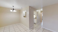 This single-floor unit offers a clean, functional layout highlighted by light-toned ceramic tile flooring that runs throughout the main living areas, reflecting natural light and contributing to a bright, airy atmosphere. The interior features a textured ceiling and neutral beige walls, creating a simple and cohesive aesthetic throughout. A decorative multi-bulb chandelier adds a subtle accent above the dining area, which connects to the rest of the living space through an open, flowing arrangement. A pass-through window between the dining area and kitchen provides convenient connectivity while maintaining a sense of defined zones. The kitchen is equipped with white cabinetry, a range, and an overhead microwave or vent hood, keeping the space practical and efficient. A hallway beyond the main living area leads further into the unit, suggesting additional private rooms. The entry exterior is equally inviting, featuring bold red doors with glass panel detailing, terracotta tile porch flooring, and decorative potted plants that add a welcoming touch. Residents also have access to a generously sized outdoor swimming pool surrounded by lush green lawn, mature palm trees, and lounge seating — offering a refreshing outdoor retreat ideal for relaxation.