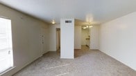 This single-floor space offers a straightforward, functional layout designed for comfortable everyday living. The main living area is finished with wall-to-wall carpet in a neutral tone, creating a warm and cohesive feel throughout. A prominent structural column positioned near the center of the space serves as a natural divider, subtly separating the entry and living zones without fully enclosing them. The textured white ceiling adds depth to the interior, while a modest ceiling fixture provides ambient lighting in the living area. The front entry door opens directly into the main space, with a hallway leading further into the unit. Adjacent to the living area, the kitchen is open and visible from the common space, featuring white cabinetry, a countertop workspace, and an elegant chandelier that adds a touch of classic charm. The kitchen's layout appears efficient and well-organized, offering ample cabinet storage. Neutral wall tones are carried throughout, lending the space a clean, airy aesthetic. An HVAC vent integrated into the central column ensures climate control is distributed effectively across the open floor plan. Overall, the space presents a tidy, move-in-ready interior with a cohesive design sensibility.