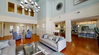 This well-appointed community clubhouse is defined by its dramatic double-height ceilings in the central gathering area, where soft blue walls, crisp white wainscoting panels, and warm hardwood flooring establish a refined yet welcoming atmosphere. An oversized chandelier anchors the main lounge, which is furnished with tufted blue accent seating, a curved sofa grouping, and glass-topped coffee tables arranged to encourage conversation. Tall, grid-paned windows flood the interior with natural light and offer views of the surrounding courtyard and greenery, while sheer drapery softens the light throughout the day.

Adjacent to the main lounge, a casual media lounge provides flat-screen televisions, built-in bar-height seating along a counter, teal booth seating, and additional lounge chairs — ideal for informal gatherings. A dedicated games room features a full-sized billiards table beneath recessed lighting and a pendant fixture, complemented by a wall-mounted television and mirrored accents.

A café-style nook with high bistro tables and teal upholstered barstools overlooks the courtyard through large windows. The catering kitchen is fully equipped with white shaker cabinetry, stainless steel appliances, a deep sink, and a coffee station, providing practical support for events and everyday use throughout the facility.