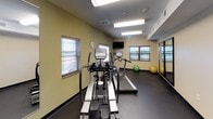 This single-level fitness room is designed to provide a functional and focused workout environment. The space features durable dark rubber flooring throughout, which is both practical and easy to maintain. Walls are finished in a combination of sage green and neutral tones, lending the room a clean, energizing atmosphere.

The cardio section is well-appointed with multiple treadmills and an elliptical trainer from the True Fitness brand, catering to a variety of aerobic exercise preferences. A wall-mounted flat-screen television is positioned for easy viewing during workouts. Stability balls are available for core training and stretching routines.

On the strength training side, the room includes a multi-function cable and weight machine alongside a flat workout bench, offering users options for a comprehensive resistance training session. A full-length mirror along one wall enhances the sense of space and allows for form checks during exercise.

The room is naturally lit through multiple windows fitted with horizontal blinds, complemented by overhead fluorescent lighting for consistent illumination. A standing fan provides additional airflow. Near the entry, a wall-mounted water fountain, paper towel dispenser, and sanitation supplies ensure cleanliness and convenience. A keyed entry door adds a layer of security to the space.