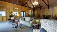 This charming single-floor space exudes rustic elegance through its masterful use of natural wood throughout. The most commanding architectural feature is the dramatic vaulted ceiling, supported by exposed wooden beams that draw the eye upward and lend a sense of grand, open volume to the interior. Warm knotty pine paneling lines the walls from floor to ceiling, creating a cohesive, cabin-inspired atmosphere that feels both cozy and spacious.

The main living area is generously proportioned, anchored by a comfortable seating arrangement that includes a classic rolled-arm sofa with decorative throw pillows and coordinating floral accent chairs. A wrought-iron and glass coffee table adds a refined, artisan touch to the sitting area. A chandelier overhead provides warm ambient lighting that complements the natural tones of the wood.

French doors with glass panes allow natural light to filter into the space while offering views of and access to the exterior. A glimpse of an adjacent area suggests additional functional spaces within the layout. Decorative elements including fresh flowers, candles, and lush potted plants add life and color to the warm, earthy palette. Overall, the space strikes a welcoming balance between rustic character and comfortable, well-appointed living.