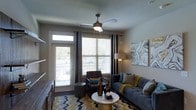 This single-floor residence offers a thoughtfully designed living space that blends comfort and contemporary style. The living area serves as the heart of the home, centered around large windows dressed with elegant dark curtain panels that allow abundant natural light to filter in while offering privacy. A sleek ceiling fan with an integrated light fixture adds both function and visual interest overhead.

The space is furnished with a plush gray sofa accented with teal and yellow throw pillows, complemented by a dark leather accent chair and a round white coffee table. A bold chevron-patterned area rug in yellow, gray, and navy anchors the seating arrangement and adds a dynamic design element to the room. Decorative accents, including bronze bottle vases and abstract wall art, contribute to the modern, curated aesthetic.

A glass-paneled entry door with a transom window leads directly to an outdoor balcony, providing an easy connection to the exterior and enhancing the sense of openness. Natural light from both the door and the large adjacent window creates a bright, welcoming atmosphere throughout. The neutral wall tones serve as a versatile backdrop, allowing the furnishings and decor to take center stage in this inviting, well-lit living space.