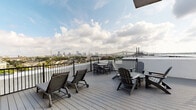 This rooftop deck in New Orleans, Louisiana, delivers an exceptional open-air experience elevated above the surrounding neighborhood. The spacious deck features durable composite wood-style decking and is enclosed by a sturdy black metal railing that ensures safety while preserving unobstructed sightlines. The standout feature of this space is its breathtaking panoramic view, encompassing the New Orleans downtown skyline, the iconic Crescent City Connection bridge spanning the Mississippi River, and the historic rooftops of the surrounding neighborhood below.

The deck is thoughtfully furnished for both relaxation and outdoor dining. Adjustable lounge chairs with cushions invite sunbathing, while Adirondack chairs and side tables create a casual seating area. A round outdoor dining table with matching chairs provides a setting for al fresco meals with a stunning city backdrop. Access to the deck is provided through a solid utility door set within stucco and exposed historic brick walls, adding an authentic New Orleans architectural character. The combination of open sky, sweeping urban views, and comfortable outdoor furnishings makes this rooftop deck a remarkable outdoor retreat in the heart of New Orleans.