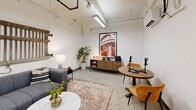This podcast studio is a thoughtfully designed single-room space that blends industrial character with modern comfort. The exposed ceiling reveals structural pipes, conduits, and track lighting, lending an authentic loft-like aesthetic to the bright, white-walled interior. Polished concrete floors anchor the space and complement the raw architectural elements above.

The room is divided into two functional zones. A lounge area features a plush gray sofa adorned with patterned throw pillows, positioned in front of a rustic reclaimed wood wall piece that serves as a striking focal point. A round marble-top coffee table and a coordinating patterned area rug complete this welcoming seating vignette, ideal for pre-recording prep or casual conversation.

The recording zone is equipped with a round wooden table, two mustard-orange accent chairs, and professional podcast microphones ready for use. A wooden credenza beneath a large-format industrial photograph displays an "ON AIR" sign and additional audio gear. A wall-mounted split-system air conditioning unit ensures climate comfort during sessions. The studio is accessed through a solid gray door and includes posted studio rules, reflecting a professional, well-managed recording environment.