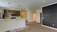 This single-floor space showcases an open-concept design that integrates the kitchen and living area into one cohesive, functional layout. The kitchen is equipped with medium-toned wood cabinetry contrasted by white lower cabinetry and a white quartz or solid-surface countertop. A subway tile backsplash adds a classic touch, while stainless steel appliances, including an over-the-range microwave, contribute to a contemporary aesthetic. A peninsula with seating overhang extends into the living space, making it ideal for casual dining or entertaining. A pendant light with a dark, industrial-style fixture hangs above the peninsula, adding a design focal point. The living area features plush carpeting, creating a warm and comfortable atmosphere. A distinctive dark brick accent wall on one side of the space adds texture and visual contrast against the otherwise neutral white walls. Recessed lighting and flush-mount ceiling fixtures provide ample illumination throughout. A hallway visible in the background leads to additional rooms, suggesting a practical and well-organized floor plan. Overall, the space balances modern finishes with inviting, comfortable elements.