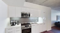 This single-floor space showcases a clean, modern aesthetic rooted in contemporary design principles. The kitchen stands as a highlight of the space, outfitted with high-gloss white flat-panel cabinetry that extends from floor to ceiling, maximizing storage while maintaining a streamlined, uncluttered appearance. The cabinetry is complemented by sleek brushed metal hardware, adding a subtle industrial touch. Stainless steel appliances, including a built-in electric range with oven, an over-the-range microwave, and a double-basin stainless steel sink, provide a fully equipped and functional cooking environment. A gray tile backsplash runs the length of the countertop, offering a neutral contrast to the bright white cabinetry and light-toned countertops. One of the most distinctive architectural features is the exposed concrete ceiling, which lends an urban, loft-inspired character to the space. This raw material juxtaposes beautifully with the polished white finishes below, creating a dynamic visual balance between industrial and modern elements. The overall ambiance is bright, crisp, and functional — ideal for those who appreciate minimalist design paired with practical, well-appointed living spaces.
