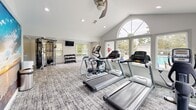 Located at 1220 Meredith Dr in Columbia, SC, this fitness center is a thoughtfully designed amenity space that offers a bright and welcoming environment for residents to stay active. The main workout area features soaring vaulted ceilings with recessed lighting and a ceiling fan, creating an airy, open atmosphere. A striking arched window on the far wall, flanked by additional large windows on multiple sides, floods the space with natural light and provides pleasant outdoor views of the surrounding pool area and greenery. The flooring is covered in a durable, patterned carpet suited for active use. Exercise equipment includes multiple treadmills, an elliptical trainer, a stationary bike, and a multi-function cable weight machine, complemented by a rack of free weights. A wall-mounted television adds entertainment during workouts, while a large scenic mural serves as a bold decorative focal point. Full-length mirrors on one wall enhance the sense of spaciousness and support strength training. A compact, dedicated business nook adjoins the main area, featuring warm hardwood-style flooring, a built-in desk with a decorative textured panel backdrop, a computer workstation, and a printer — offering a convenient spot for residents to handle everyday tasks.