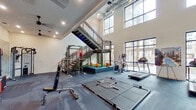 This expansive two-story fitness center is designed to support a full range of workout disciplines within a modern, well-appointed environment. The ground floor opens to soaring double-height ceilings and is flooded with natural light through large, industrial-style floor-to-ceiling windows that provide views of the surrounding outdoor areas. This level is equipped with multiple treadmills, an elliptical trainer, a stair climber, power racks with free weights, a dumbbell rack, plyo boxes, and dedicated turf and mat zones for functional and strength training. A decorative tiled accent wall adds visual interest near the entry point on this floor. A striking open-riser metal staircase with mesh railings connects the two levels, serving as both a functional and architectural centerpiece. The upper floor features a spacious open layout with patterned carpet flooring, exposed ductwork overhead, and an array of sleek pendant lighting. Floor-to-ceiling mirrors line the walls, amplifying the sense of space and supporting form-focused training. This level houses additional strength and conditioning equipment, battle ropes, medicine balls, kettlebells, resistance machines, and a mounted flat-screen display. Together, both floors deliver a comprehensive, professional-grade fitness experience within a thoughtfully designed space.