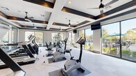 This fitness center is a thoughtfully designed, single-floor facility that offers a comprehensive workout environment. The space is anchored by a striking coffered ceiling with dark wood trim accents, recessed lighting, and multiple ceiling fans that keep the atmosphere comfortable during exercise. Large floor-to-ceiling sliding glass windows and doors line one side of the space, flooding the interior with natural light and providing direct views of an outdoor pool and patio area, creating an open, airy ambiance. The main workout floor is finished with a neutral, grid-patterned carpet tile that provides both durability and comfort underfoot. It is furnished with a full suite of cardio equipment, including upright stationary bikes, recumbent bikes, elliptical machines, and treadmills, complemented by a multi-station cable machine for strength training. Large framed mirrors along one wall visually expand the space and allow users to monitor their form. A dedicated free-motion room adjacent to the main floor offers open space for stretching and functional training, equipped with medicine balls and wall-mounted art. Near the entrance, built-in cubbies and storage provide organizational convenience. Wall-mounted televisions are positioned throughout for entertainment during workouts.