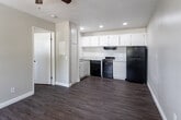 Located at 1401 E Santo Antonio Dr in Colton, California, this single-floor unit offers a clean, functional layout finished with warm wood-look vinyl flooring that runs consistently throughout the space. The open living area welcomes natural light through a window fitted with blinds and is equipped with a ceiling fan, creating a comfortable, well-ventilated environment.

The kitchen is efficiently designed in an L-shaped configuration, featuring white cabinetry that extends to the upper and lower sections of the walls, providing ample storage. Light-toned countertops contrast neatly with the black appliance suite, which includes a gas range, dishwasher, refrigerator, and a built-in range hood. Recessed lighting illuminates the kitchen workspace effectively.

Adjacent to the main living space, the bedroom offers a calm, neutral atmosphere with soft warm-toned walls, a window with natural light, and a wall-mounted air conditioning unit for climate control. The bedroom provides direct access to a full bathroom, which includes a shower-tub combination and toilet.

Throughout the unit, clean white baseboards and neutral wall tones contribute to a bright, well-maintained appearance. The straightforward single-floor layout ensures easy navigation between all areas, making this a practical and comfortable living space.