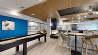 This expansive single-floor amenity space offers a thoughtfully designed open-concept layout that seamlessly blends entertainment, dining, and lounge areas into one cohesive environment. Light wood-look tile flooring flows throughout, anchoring the modern aesthetic defined by a mix of warm wood accents, crisp white walls, and bold pops of cobalt blue.

The gourmet kitchen is a standout feature, showcasing a striking marble-look waterfall island with bar seating, dark cabinetry, stainless steel appliances, and a geometric white tile backsplash. Large windows flood the space with natural light and offer views to the exterior. Pendant lighting adds a refined finishing touch.

The living and lounge area centers around a dramatic double-sided gas fireplace framed in dark stone, paired with a mounted television and comfortable seating arrangements. A dedicated game zone includes both a full-sized billiards table and a shuffleboard table, perfect for social gatherings.

Multiple dining configurations are available, including an oval dining table with seating for eight, as well as casual bar-height seating along a central counter beneath globe pendant chandeliers. Wood-paneled ceiling details and varied ceiling heights add architectural dimension throughout, creating distinct zones within the open floor plan.