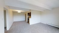 This single-floor space offers a clean, functional layout centered around an open-concept living area. The generously sized main room is finished with soft, neutral-toned wall-to-wall carpeting that adds warmth and comfort underfoot, while crisp white walls and a textured ceiling contribute to a bright and airy atmosphere. A structural beam subtly defines the transition between the living space and the kitchen area, adding architectural character without disrupting the open feel.

The kitchen is semi-enclosed, framed by a half-wall pass-through that maintains visual connectivity with the living area. It features stainless steel appliances, including a range and overhead microwave, complemented by dark wood cabinetry that provides a warm contrast to the lighter tones throughout the rest of the space. A flush-mounted ceiling light illuminates the kitchen, ensuring a well-lit workspace.

The overall design is straightforward and practical, with a neutral color palette that makes it easy to personalize. The layout flows naturally from the main living space into the kitchen, making it well-suited for everyday living. The combination of plush carpeting, clean lines, and functional kitchen amenities creates a comfortable and inviting environment.