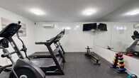 Located at 585 Ellsworth St in Bridgeport, Connecticut, this fitness room offers a compact yet efficiently organized exercise space designed to support a well-rounded workout routine. The room features clean white walls, white trim, and durable rubber flooring in a speckled gray finish, providing a practical and polished aesthetic throughout.

The space is equipped with a treadmill, elliptical trainer, stationary upright bike, and a rowing machine, offering a comprehensive range of cardio options. A set of dumbbells and a medicine ball round out the available fitness equipment for supplemental strength and conditioning work. A wall-mounted flat-screen television is positioned for easy viewing during workouts.

A large wall mirror spans one side of the room, enhancing the sense of openness and allowing users to monitor their form. Flush-mounted ceiling lights provide bright, even illumination throughout. A wall-mounted split air conditioning unit ensures climate comfort during use.

Practical amenities include a hand sanitizer dispenser, a waste receptacle, and clearly marked emergency exits, reflecting attention to hygiene and safety. The room is accessible via two doors, supporting smooth entry and exit flow. Motivational artwork accents the walls, adding a touch of energy to the overall environment.
