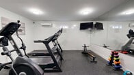 Located at 585 Ellsworth St in Bridgeport, Connecticut, this fitness room offers a compact yet efficiently organized exercise space designed to support a well-rounded workout routine. The room features clean white walls, white trim, and durable rubber flooring in a speckled gray finish, providing a practical and polished aesthetic throughout.

The space is equipped with a treadmill, elliptical trainer, stationary upright bike, and a rowing machine, offering a comprehensive range of cardio options. A set of dumbbells and a medicine ball round out the available fitness equipment for supplemental strength and conditioning work. A wall-mounted flat-screen television is positioned for easy viewing during workouts.

A large wall mirror spans one side of the room, enhancing the sense of openness and allowing users to monitor their form. Flush-mounted ceiling lights provide bright, even illumination throughout. A wall-mounted split air conditioning unit ensures climate comfort during use.

Practical amenities include a hand sanitizer dispenser, a waste receptacle, and clearly marked emergency exits, reflecting attention to hygiene and safety. The room is accessible via two doors, supporting smooth entry and exit flow. Motivational artwork accents the walls, adding a touch of energy to the overall environment.