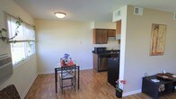 This single-floor unit offers a practical and efficiently designed open-concept layout, where the dining and kitchen areas flow naturally into one another. The space is finished with warm hardwood-style flooring that extends throughout, lending a cohesive and inviting feel to the interior.

The kitchen is tucked neatly into a defined nook, featuring medium-toned wood cabinetry with ample upper storage, dark countertops, a stainless steel dishwasher, a black gas range, and a microwave. A recessed partition wall clearly delineates the kitchen from the dining area while maintaining the open feel of the space. The kitchen also includes a sink positioned beneath upper cabinetry, maximizing functionality within a compact footprint.

Adjacent to the kitchen, the dining area comfortably accommodates a small bistro-style table and chairs, making it an ideal spot for casual meals. A potted plant adds a touch of greenery to the space. The walls are painted in a soft, neutral yellow tone, creating a bright and welcoming atmosphere. A flush-mount ceiling light provides overhead illumination, and the HVAC vents integrated into the upper wall ensure year-round comfort. The overall design is clean, functional, and well-suited for everyday living.
