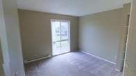 This single-floor space offers a clean and straightforward layout designed for comfortable everyday living. The interior is finished with wall-to-wall carpeting in a neutral tone, providing a soft and cohesive feel throughout. Walls are painted in a warm greige hue, creating a calm and understated ambiance that complements a variety of furnishing styles. A textured white ceiling adds subtle visual interest overhead. The space benefits from a large double-hung window that fills the room with generous natural light and frames a pleasant view of lush green outdoor foliage, contributing to a bright and refreshing atmosphere. White trim and baseboards provide clean contrast against the neutral walls, lending the space a tidy, well-maintained appearance. A baseboard heating unit runs along the lower portion of the exterior wall, ensuring efficient and consistent warmth throughout the colder months. The overall character of the space is practical and inviting, with a simple design palette that offers a versatile backdrop ready to be personalized to suit individual preferences.