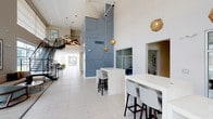 The clubhouse lobby is a contemporary, well-appointed common space designed to create a welcoming first impression. The most striking architectural feature is the soaring double-height ceiling, which lends the space an open, airy quality and amplifies the sense of scale throughout. A bold, floor-to-ceiling geometric accent wall in muted blue-grey serves as the focal point of the lobby, adding a distinctive artistic element to the overall design.

Light-toned large-format tile flooring runs throughout, contributing to the bright and clean aesthetic. Overhead, sculptural gold geodesic pendant lights are suspended at varying heights, providing warm ambient lighting and serving as decorative focal points against the white ceiling.

White high-top tables with modern bar-height seating are strategically positioned throughout the space, offering residents casual areas to gather or work. A staircase with sleek black metal railings is visible to one side, adding to the architectural interest of the lobby.

Adjacent rooms are accessible through framed openings, hinting at additional amenity spaces beyond. Large windows near the entrance allow natural light to filter in, enhancing the lobby's bright, polished, and modern character.