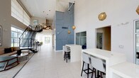 The clubhouse lobby is a contemporary, well-appointed common space designed to create a welcoming first impression. The most striking architectural feature is the soaring double-height ceiling, which lends the space an open, airy quality and amplifies the sense of scale throughout. A bold, floor-to-ceiling geometric accent wall in muted blue-grey serves as the focal point of the lobby, adding a distinctive artistic element to the overall design.

Light-toned large-format tile flooring runs throughout, contributing to the bright and clean aesthetic. Overhead, sculptural gold geodesic pendant lights are suspended at varying heights, providing warm ambient lighting and serving as decorative focal points against the white ceiling.

White high-top tables with modern bar-height seating are strategically positioned throughout the space, offering residents casual areas to gather or work. A staircase with sleek black metal railings is visible to one side, adding to the architectural interest of the lobby.

Adjacent rooms are accessible through framed openings, hinting at additional amenity spaces beyond. Large windows near the entrance allow natural light to filter in, enhancing the lobby's bright, polished, and modern character.