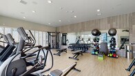Located at 160 S Ventura Ave in Ventura, California, this single-floor fitness center is a modern, thoughtfully designed facility built to support a comprehensive workout experience. The space features light hardwood-style flooring throughout, recessed lighting in the smooth ceilings, and neutral-toned walls accented by warm wood paneling that lend a contemporary, clean aesthetic.

The main workout area is generously proportioned and houses an extensive selection of equipment, including treadmills, stair climbers, elliptical machines, rowing machines, and a multi-station cable machine for strength training. A full rack of dumbbells lines a mirrored wall, which visually expands the space and makes it easy to monitor form during exercise. Additional accessories such as medicine balls, resistance tools, and stability balls are neatly organized and readily accessible.

Floor-to-ceiling glass windows and sliding doors flood the space with abundant natural light and offer direct views of the outdoor pool area, creating a bright and energizing atmosphere. A dedicated area near the pool access includes a Peloton spin bike with an integrated screen, ideal for immersive cycling sessions. The seamless indoor-outdoor connection, with palm tree views visible beyond the glass, enhances the overall California lifestyle ambiance of the facility.