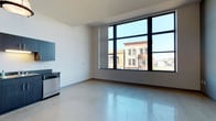 This contemporary urban unit offers a thoughtfully designed single-floor layout that blends modern finishes with an open, airy atmosphere. Polished concrete floors extend throughout the main living areas, reflecting natural light and lending an industrial-chic character to the space. Expansive floor-to-ceiling windows with sleek black frames dominate the living area, flooding the interior with abundant natural light and framing sweeping views of the surrounding cityscape and historic neighboring buildings.

The open-concept kitchen is well-equipped with a full suite of stainless steel appliances, including a range, microwave, refrigerator, and dishwasher. Dark navy cabinetry with matching dark granite countertops adds a rich contrast to the bright, white walls. Industrial-style pendant lighting above the kitchen enhances both functionality and aesthetic appeal.

The bedroom benefits from the same generous window presence found in the living area, offering continued city views and natural light. The bathroom features a dark granite vanity countertop with an undermount sink, warm wood-toned cabinetry and doors, tile flooring, and a large mirror. A walk-in closet with built-in shelving provides practical storage. Wall sconces throughout the unit add warm accent lighting, completing the modern yet inviting ambiance of this urban residence.