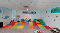 Located at 153-30 89th Avenue in Jamaica, NY, this single-floor children's space is thoughtfully designed around an immersive underwater ocean theme. The facility is divided into distinct activity zones that work together to create an engaging and welcoming environment for children. The play area features a bright, multicolored interlocking foam tile floor that provides a safe, cushioned surface for active play. Soft foam building blocks, a child-sized basketball hoop, and a small slide offer hands-on entertainment, while a panoramic porthole-style mural depicting a vibrant coral reef serves as a charming focal point on the light blue walls. Whimsical marine creature decals — including sharks, fish, and an octopus — are distributed across the walls, reinforcing the undersea atmosphere throughout. A separate lounge zone provides a more relaxed setting, furnished with a teal sofa and a wall-mounted flat-screen television, alongside an interactive bead maze activity table. Shark-shaped bean bag seats and plush floor cushions add playful comfort to this area. Large windows fitted with colorful ocean-scene window treatments allow natural light to filter in. The speckled terrazzo-style flooring in the lounge zone complements the drop tile ceiling with recessed lighting. Secure glass-paneled entry doors connect the space to the broader facility.