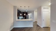 This single-floor residence offers a thoughtfully designed open-concept layout that seamlessly connects the kitchen and living areas, creating an inviting and functional living environment. The kitchen is a standout feature, showcasing rich dark cabinetry that contrasts beautifully against light gray walls and a classic subway tile backsplash. A peninsula with a granite countertop serves as both a prep space and a casual dining area, illuminated by a trio of sleek pendant lights that add a modern, polished touch. Stainless steel appliances, including a refrigerator and range, complete the kitchen's contemporary aesthetic. The flooring throughout is a warm, dark hardwood-style material that adds depth and sophistication to the space. Neutral gray walls provide a clean, versatile backdrop that complements the overall modern design. A glimpse into an adjacent room — likely a laundry or utility area — hints at the functional layout of the residence. Generous natural light fills the space, enhancing its open and airy feel. The overall ambiance is clean, contemporary, and well-appointed, making it an ideal space for both everyday living and entertaining.