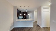 This single-floor residence offers a thoughtfully designed open-concept layout that seamlessly connects the kitchen and living areas, creating an inviting and functional living environment. The kitchen is a standout feature, showcasing rich dark cabinetry that contrasts beautifully against light gray walls and a classic subway tile backsplash. A peninsula with a granite countertop serves as both a prep space and a casual dining area, illuminated by a trio of sleek pendant lights that add a modern, polished touch. Stainless steel appliances, including a refrigerator and range, complete the kitchen's contemporary aesthetic. The flooring throughout is a warm, dark hardwood-style material that adds depth and sophistication to the space. Neutral gray walls provide a clean, versatile backdrop that complements the overall modern design. A glimpse into an adjacent room — likely a laundry or utility area — hints at the functional layout of the residence. Generous natural light fills the space, enhancing its open and airy feel. The overall ambiance is clean, contemporary, and well-appointed, making it an ideal space for both everyday living and entertaining.