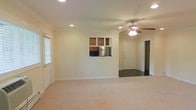 This single-floor residence offers a well-organized and functional layout designed for comfortable everyday living. The main living area is expansive and open, finished with plush wall-to-wall carpeting that adds warmth and softness underfoot. Recessed lighting is installed throughout the ceiling, providing even, ambient illumination across the space, while a ceiling fan with an integrated light fixture adds both practicality and a classic touch to the room.

A standout architectural feature is the pass-through window that connects the living area to the kitchen, allowing natural sightlines into the culinary space. Through this opening, warm wood-toned cabinetry and a stainless steel refrigerator are visible, hinting at a well-appointed kitchen beyond. The transition between the carpeted living space and the tiled entryway is clearly defined, with a front entry door leading into a foyer area. Adjacent to the entry, a closet with open shelving provides convenient storage. Crown molding along the ceiling adds a refined, traditional detail to the overall aesthetic. A window with horizontal blinds on the side wall allows for natural light and ventilation, completing this bright and inviting single-level home.