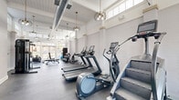 Located at 3100 Southgate Drive in Alexandria, VA, this single-floor fitness center offers a thoughtfully designed space for a comprehensive workout experience. The main exercise room is a standout, boasting soaring ceilings with exposed ductwork and elegant globe pendant lighting that adds a stylish touch to the industrial-chic aesthetic. White-painted brick walls and large glass doors flood the space with natural light and provide outdoor views, creating an open, energizing atmosphere. The rubber-floored workout area is well-appointed with a full range of cardio equipment — including treadmills, ellipticals, stair climbers, and stationary bikes — alongside rowing machines, kettlebells, dumbbells, and a weight bench for strength training. A separate, tile-floored movement studio features full-length mirrored walls, recessed lighting, and a drop ceiling, making it ideal for stretching, yoga, or group fitness. Medicine balls and accessories are neatly organized on dedicated racks. The hallway connecting these areas is clean and functional, with neutral tones and white panel doors that maintain a cohesive, polished design throughout. A water fountain station serves the main workout area, rounding out the practical amenities of this well-rounded facility.