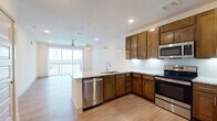 This well-designed single-floor residence offers a thoughtfully laid-out open-concept floor plan that seamlessly connects the kitchen and living areas. The kitchen serves as the heart of the home, showcasing rich dark-wood cabinetry, granite countertops, a subway tile backsplash, and a full suite of stainless steel appliances including a range, microwave, dishwasher, and refrigerator. A peninsula with an undermount sink and pendant lighting above provides additional prep space and a casual dining option. Light wood-style flooring runs throughout the main living area, complementing the neutral walls and recessed lighting. Large floor-to-ceiling windows flood the living space with natural light and offer elevated city views, while a ceiling fan adds comfort overhead. The bedroom continues the bright, airy aesthetic with plush carpeting, a ceiling fan, and a window framing urban views. The bathroom features consistent dark-wood cabinetry with granite countertops, wood-look flooring, and a tiled soaking tub and shower surround. A spacious walk-in closet with built-in shelving and hanging rods provides ample storage. Throughout the unit, consistent finishes and clean lines create a cohesive, modern living environment.