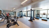Located at 37 8th Ave in Oakland, CA, this well-appointed fitness center offers a generously sized, single-floor layout designed to accommodate a full range of workout activities. The open floor plan maximizes usability, with durable rubber and carpet-tile flooring delineating distinct exercise zones throughout the space.

The cardio section is prominently equipped with multiple treadmills, elliptical trainers, and stationary cycling bikes, each outfitted with individual digital screens for an engaging workout experience. A rowing machine rounds out the cardio offerings, providing users with a comprehensive selection of aerobic equipment.

The strength training area features cable machine systems and free weight benches, offering versatile options for resistance-based exercise. Large wall-mounted mirrors run along key sections of the facility, enhancing the sense of openness while allowing users to monitor their form.

Overhead, recessed linear LED lighting and exposed ductwork add a clean, modern industrial aesthetic to the space. Natural light filters in through glass-paneled doors and windows along one wall, providing a connection to the outdoors. A wall-mounted display screen serves as an informational hub within the facility. The overall ambiance is bright, functional, and professionally designed, making this fitness center a well-equipped resource for residents or members in the Oakland area.
