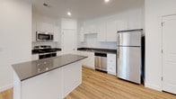 This well-designed single-floor residence offers a thoughtfully laid-out floor plan that balances functionality with modern aesthetics. The open living area greets you with light-toned hardwood-style flooring, recessed lighting, and crisp white walls that create a bright, welcoming atmosphere.

The kitchen is a standout feature, outfitted with white shaker-style cabinetry, dark quartz countertops, and a full suite of stainless steel appliances including a refrigerator, range, microwave, and dishwasher. A central island provides additional prep space and a natural gathering point, while the light flooring ties the space together seamlessly.

The bedroom is carpeted for comfort and includes a generously sized walk-in closet fitted with dual hanging rods and shelving, offering ample storage for any wardrobe. A window allows natural light to fill the room.

The full bathroom is clean and contemporary, featuring white cabinetry with a dark countertop vanity, an undermount sink, and a tub-shower combination with built-in shelving. Light wood-style flooring extends throughout, maintaining a cohesive design.

A dedicated laundry room houses a full-size top-loading washer and dryer, adding significant everyday convenience. The unit's consistent use of neutral tones and modern finishes throughout creates a polished, move-in-ready environment.